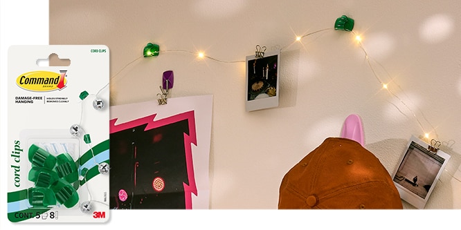 Command™ Small Cord Clips on a wall with string lights, polaroid photos, and a poster. A brown cap is hanging below the display.
