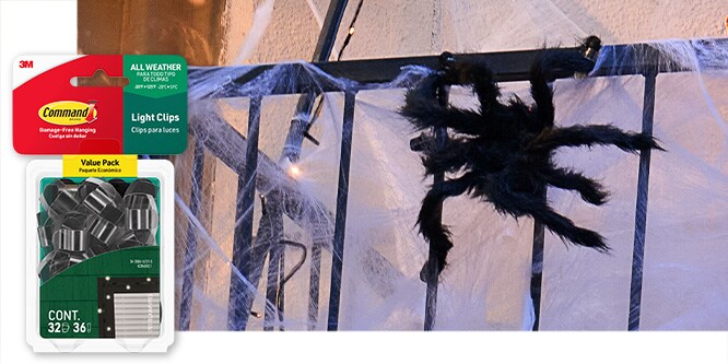 Command™ Black Outdoor Light Clips holding a spider and web decoration on a balcony railing.  Product in package in lower left corner.
