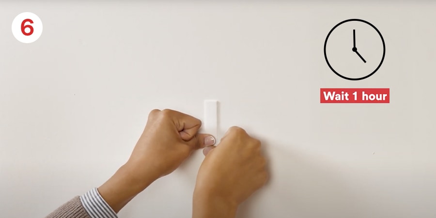 Hands press a hook onto a wall while a clock icon indicates to wait 1 hour before hanging frame.