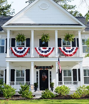 For additional patriotic decoration, place a large American flag near the front porch by placing it in a flag holder, hang large patriotic bunting flags by securing with string tied around porch railings and hang fern baskets from pre-installed hooks
