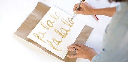 Une femme qui peinture « Fa la la la » en or sur un papier blanc.
