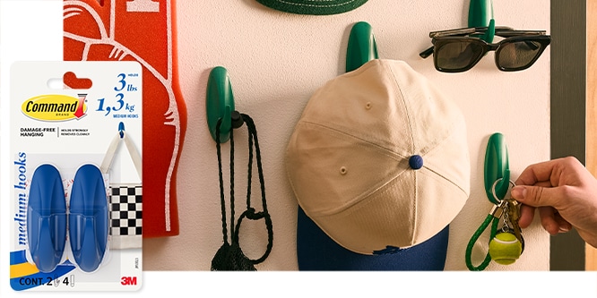 Command™ Medium Hooks on a wall holding a cap, sunglasses, and keys. Packaging shown on the left.
