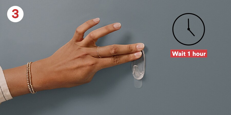Hand pressing a clear adhesive hook on a wall with instructions to wait one hour. Overlayed text: 