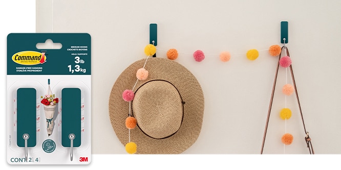 Command™ Medium Wire Hooks on a wall holding a hat, and a handbag with a string of colorful pompoms.
