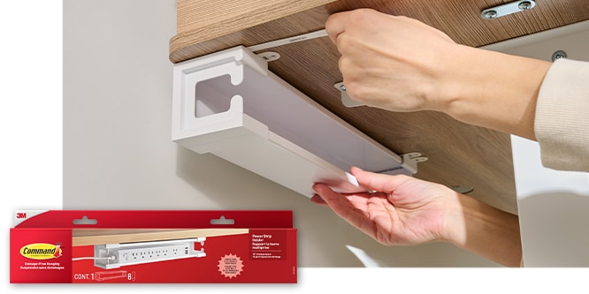 Hands securing a Command™ Power Strip holder to the underside of a desk. Product in package in lower left corner.
