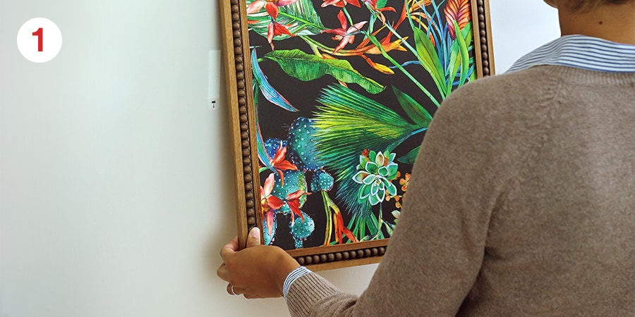 Person removing a framed tropical-themed artwork from a wall, with vibrant green leaves and flowers.