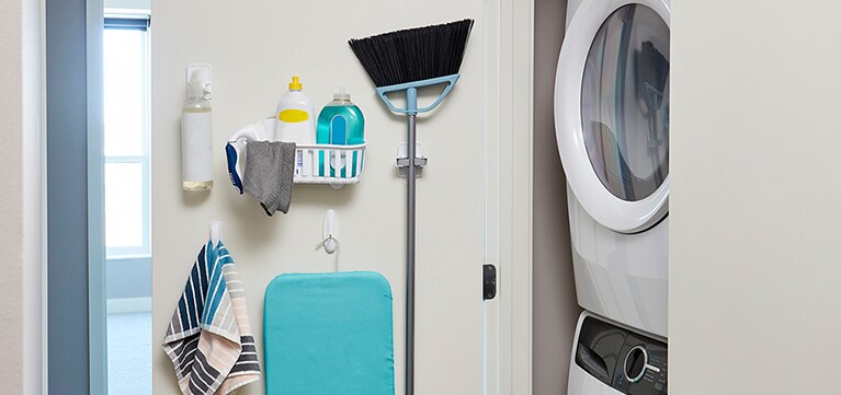 Un balai, une serviette et divers produits de nettoyage accrochés à une porte de buanderie, fixés à l’aide de crochets et d’outils Command(MC).
