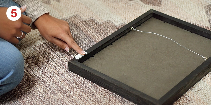 A hand presses stabilizing strips to the back of a frame on a patterned carpet.
