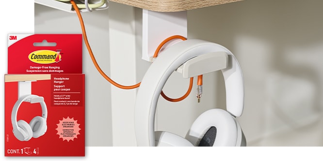 Command™ Headphone Hanger hanging a pair of headphones with an orange cord, from the underside of a desk. Product in package in lower left corner.
